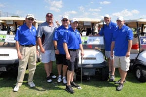 Six men posing for a picture on the golf course after playing golf with golf carts in the background.